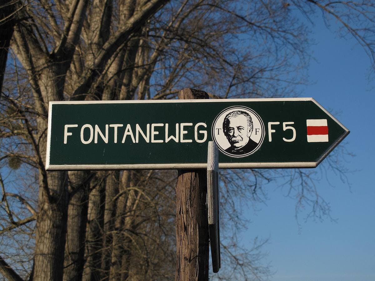 A dark green trail sign for the Fontaneweg F5 with Theodor Fontane’s portrait, against winter branches