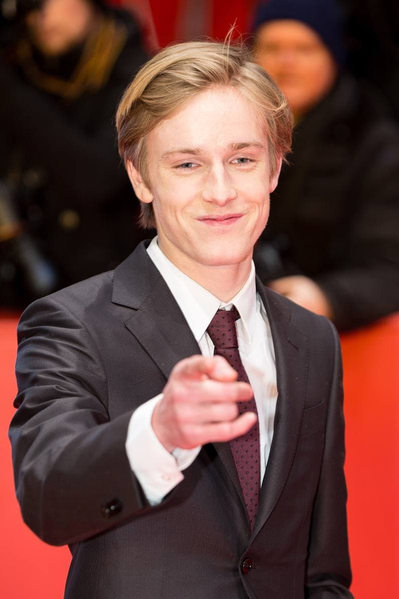 Louis Hofmann in a dark suit and tie on the red carpet, smiling and pointing