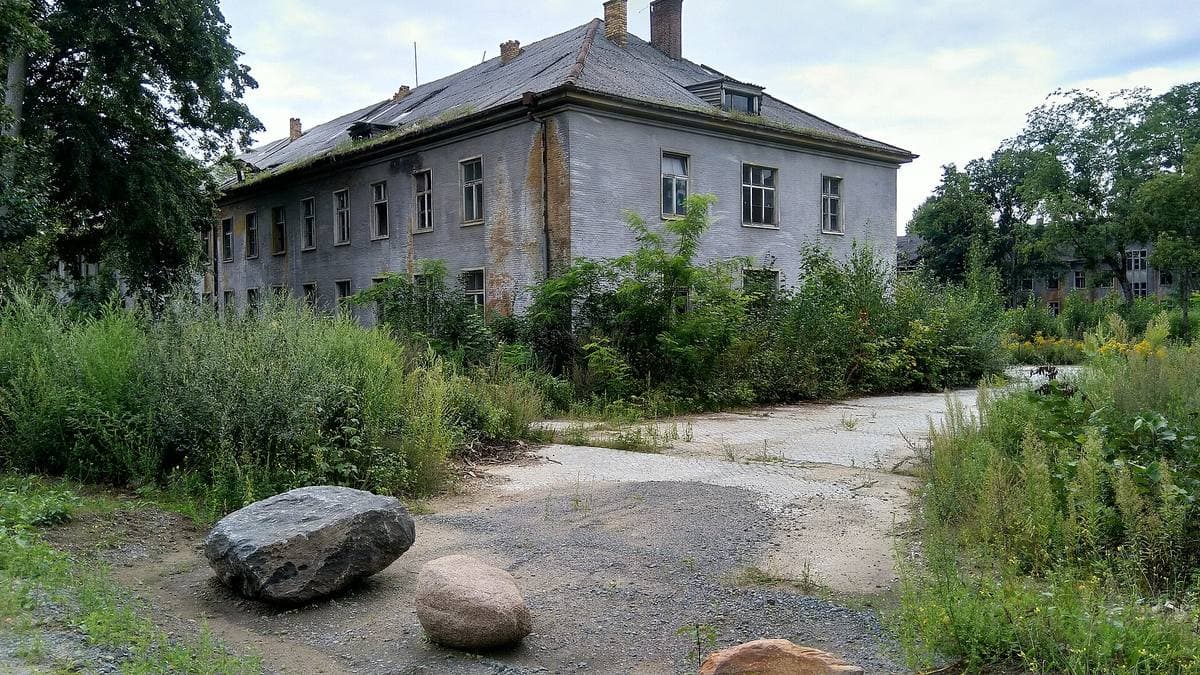 A grey two-storey barracks block behind long grass, a cracked track and boulders in front