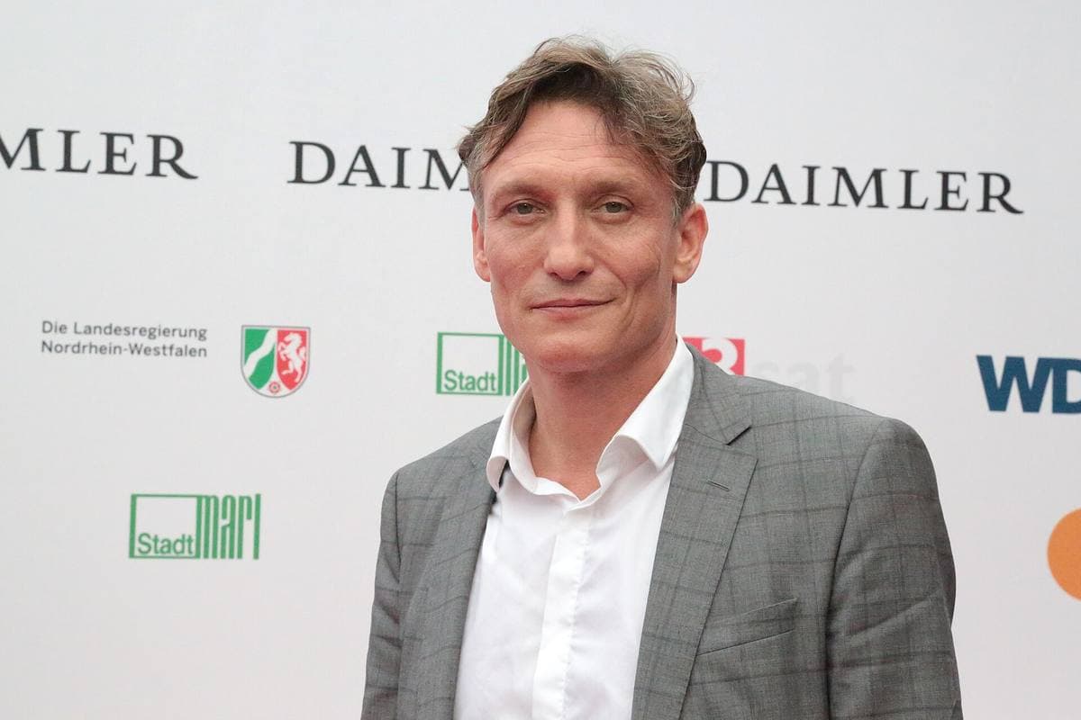 Oliver Masucci in a grey checked jacket and open white shirt