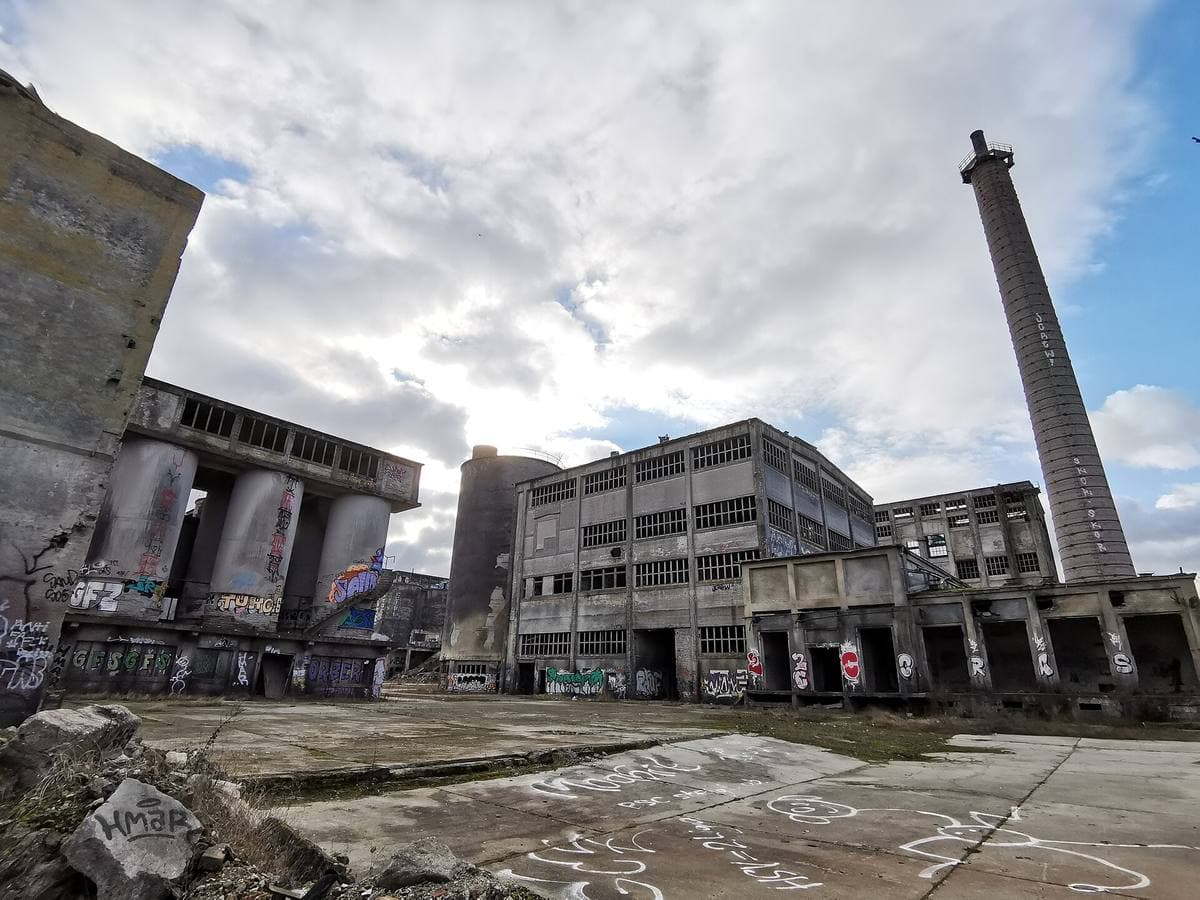 Gutted concrete factory halls and a tall chimney over a broken yard covered in graffiti
