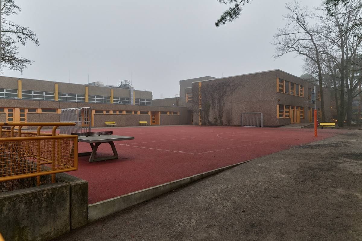 Flat-roofed brick school buildings round a red sports court on a misty day
