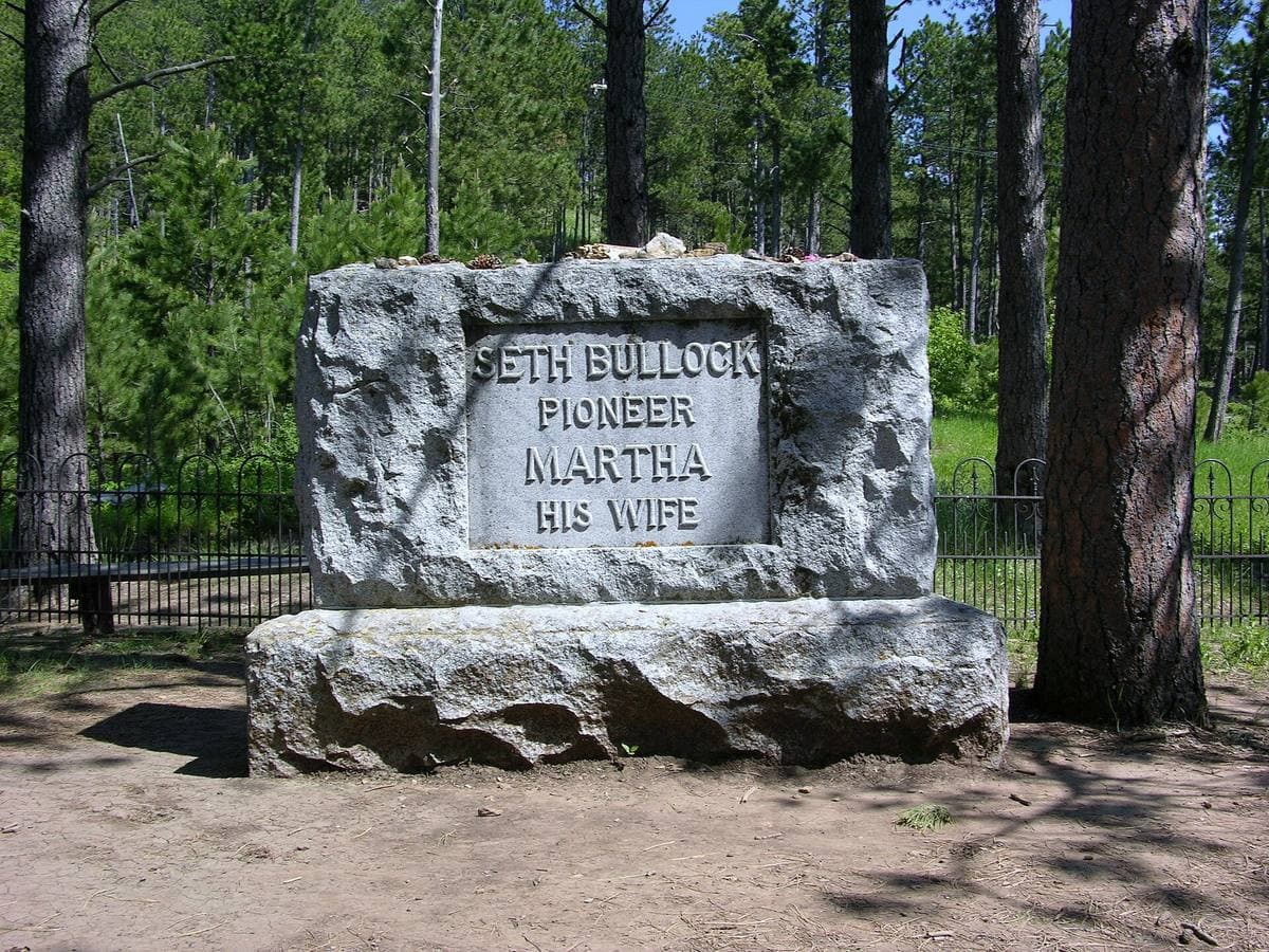 A rough stone grave marker inside a small iron fence among pines