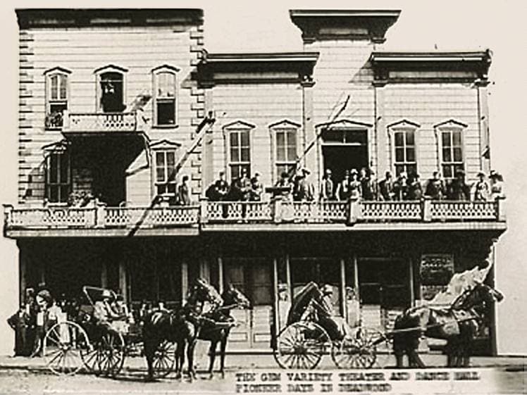 A two-storey wooden theatre front with a long balcony and horses and carriages outside