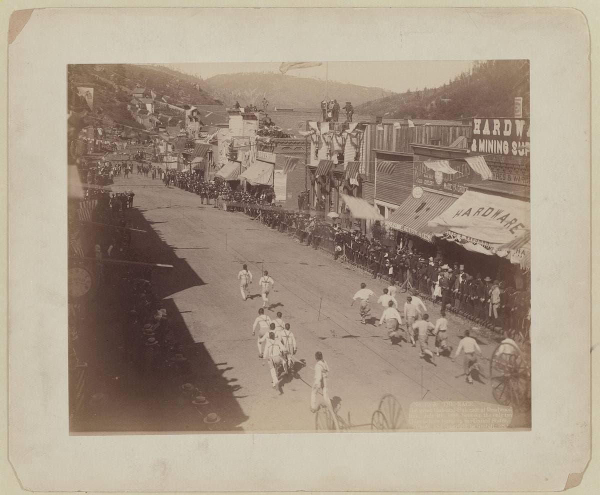 Crowds and running men on a wide Main Street lined with brick buildings