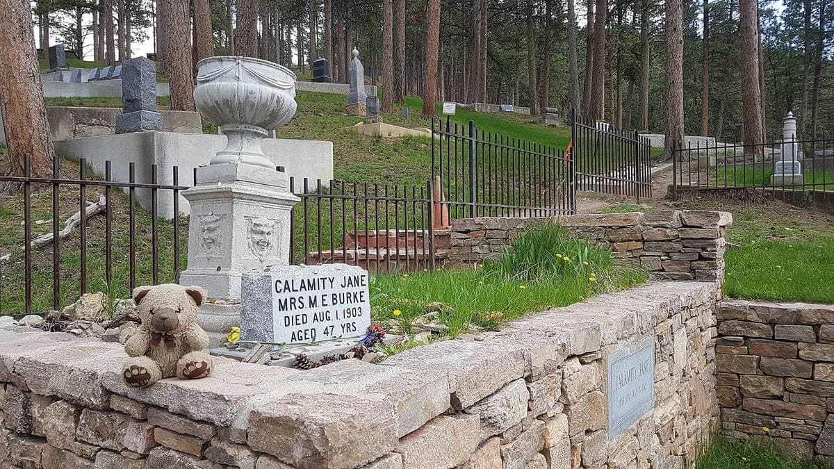 A railed grave plot with a stone marker and flowers among pines