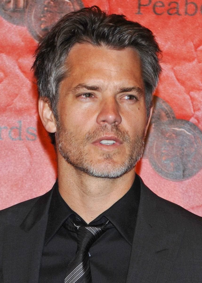 Timothy Olyphant in a dark suit and tie against a red wall