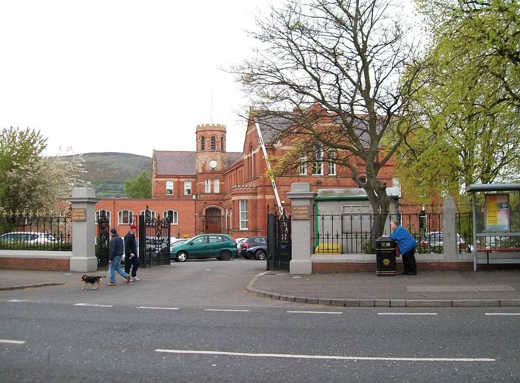 Red-brick college buildings behind railings and trees on a main road