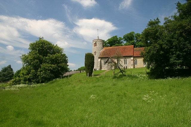 A small church with a round flint tower among trees above a green meadow