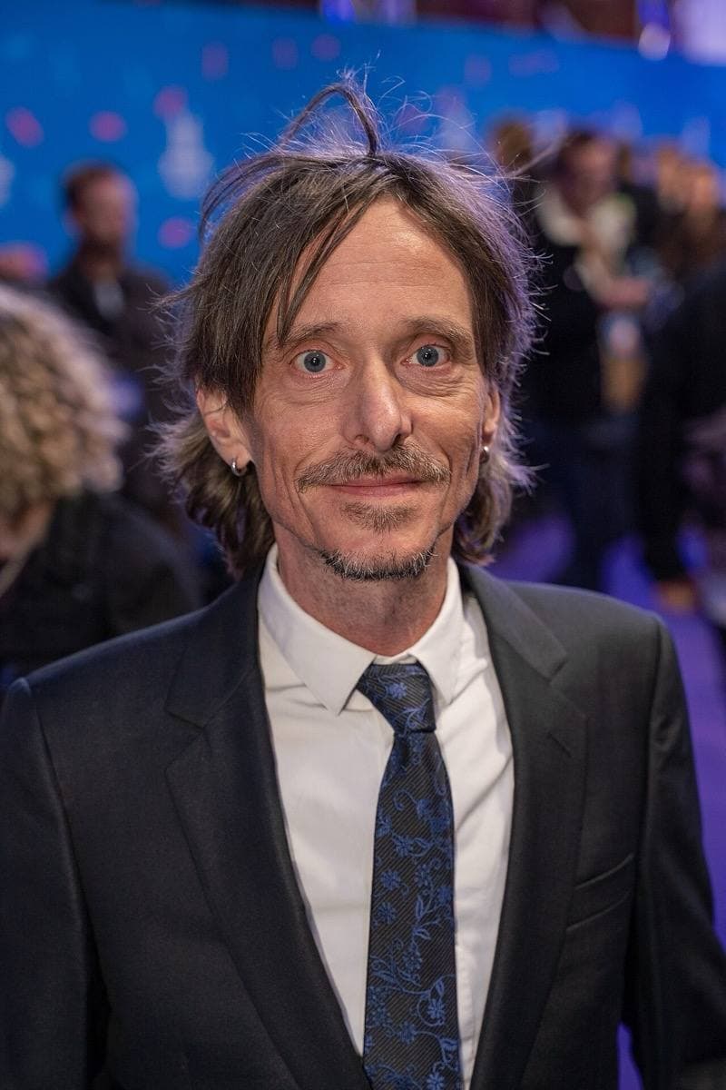 A thin man with long tousled hair and a moustache in a dark suit and tie, smiling