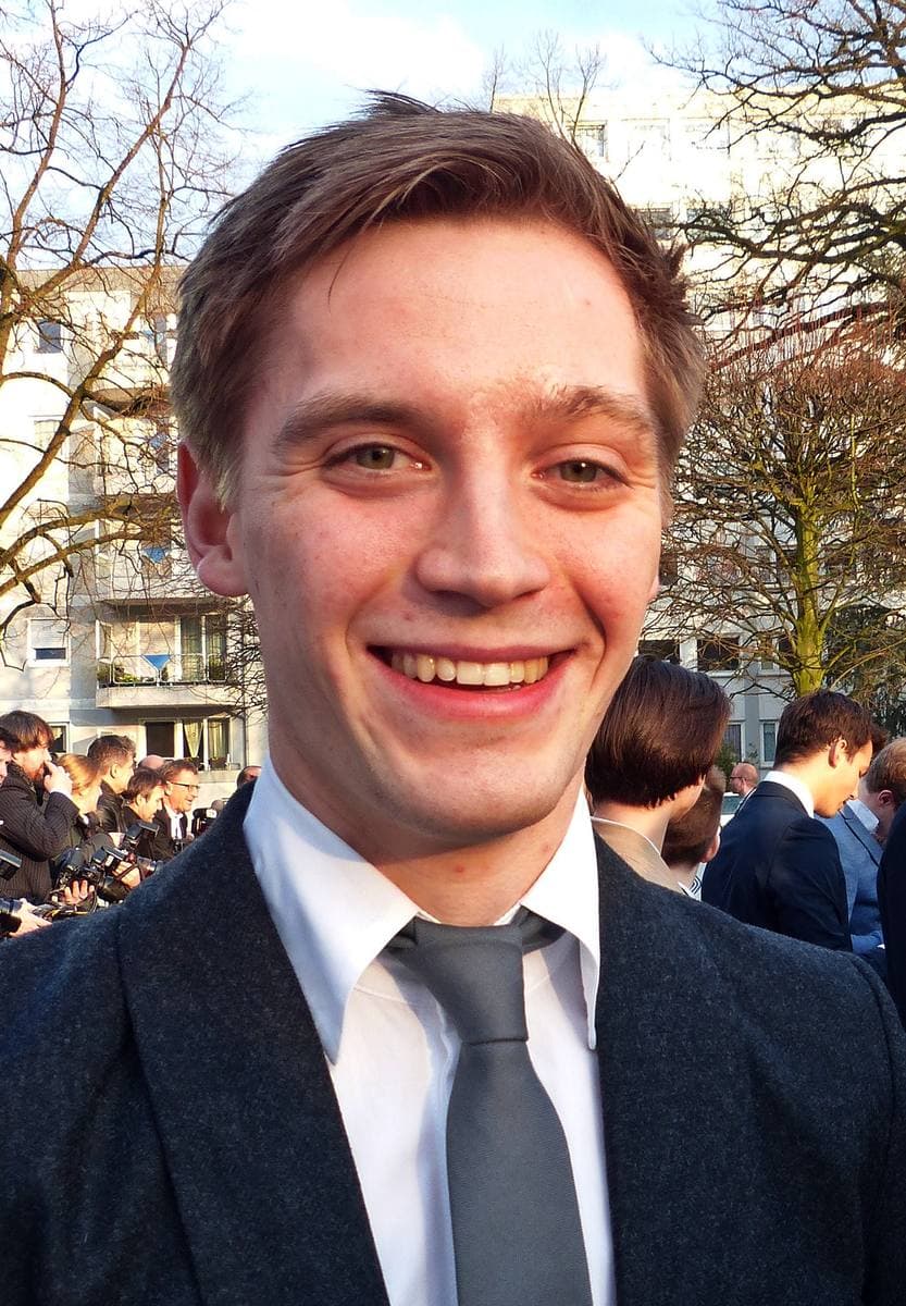 Jonas Nay in a dark suit and tie, smiling, in front of a sponsor wall