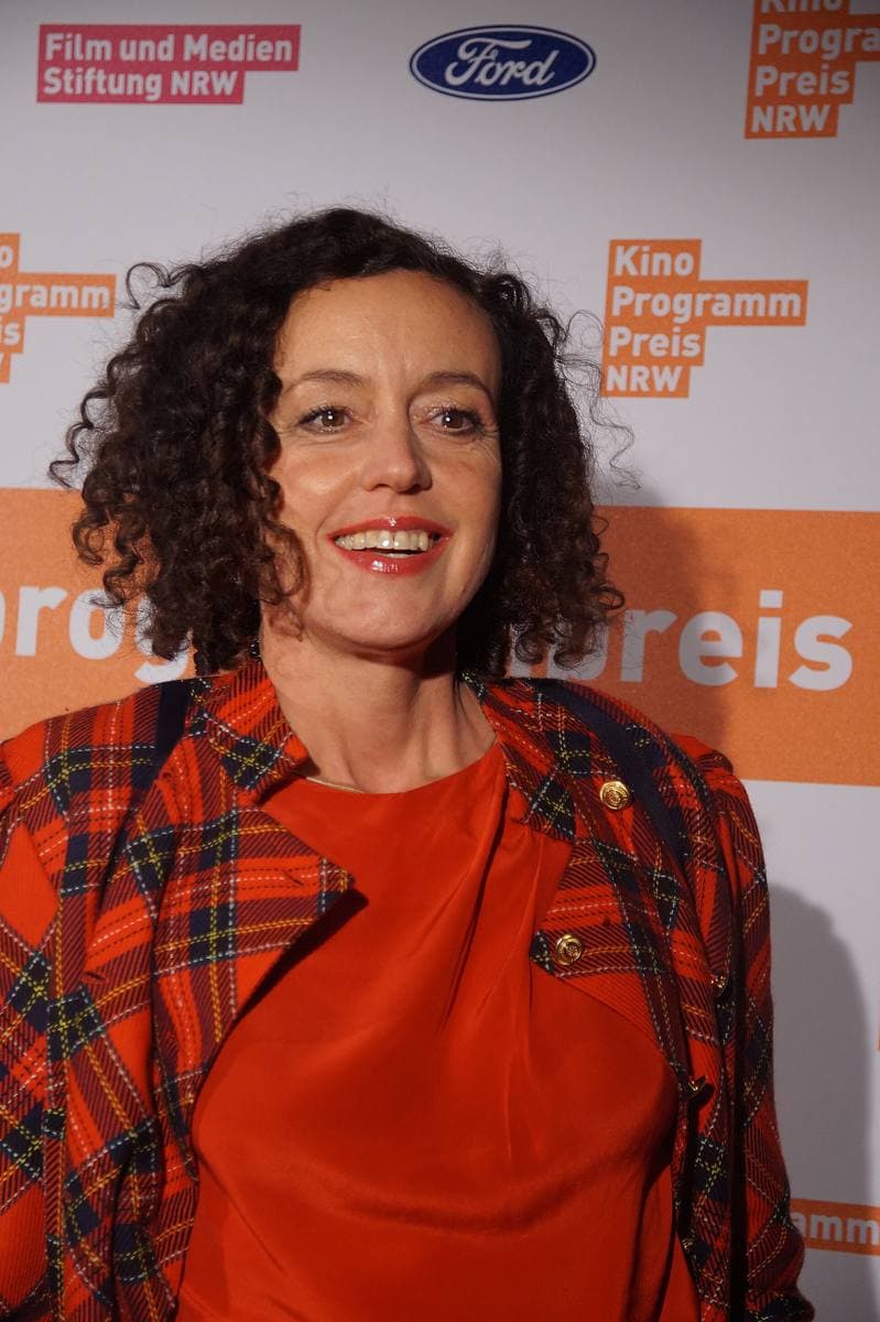 Maria Schrader with curly dark hair and a red checked coat, smiling