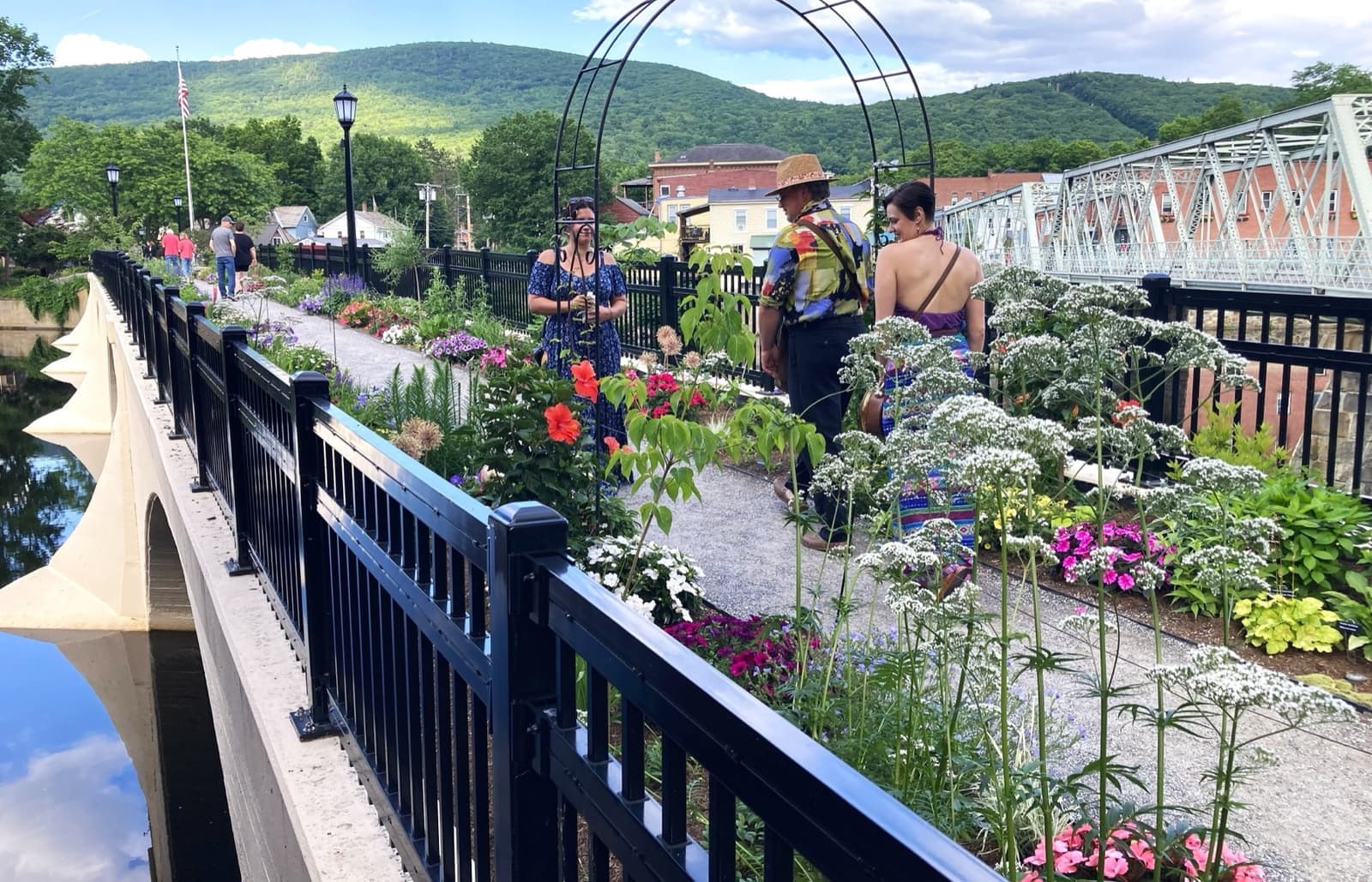 Visitors on a flower-lined footbridge beside an iron truss bridge