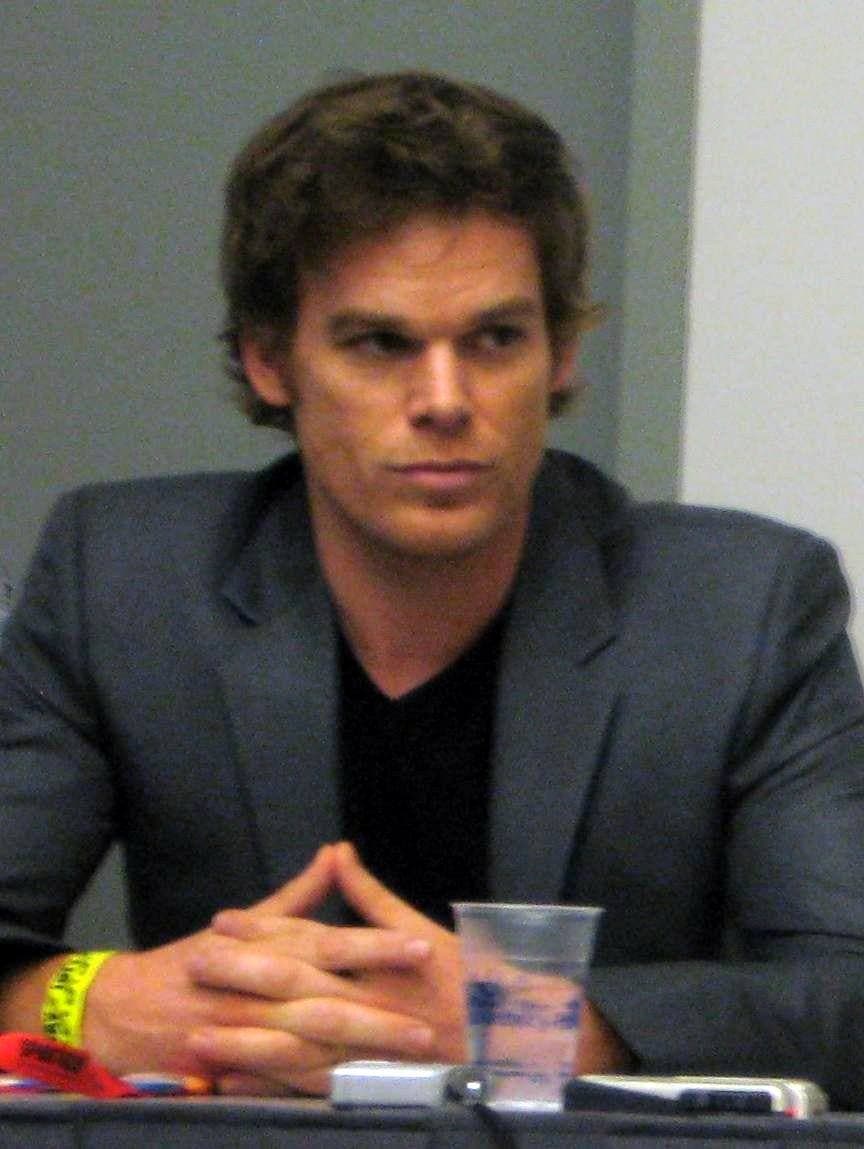 Michael C. Hall in a grey jacket, hands clasped on a table, looking past the camera