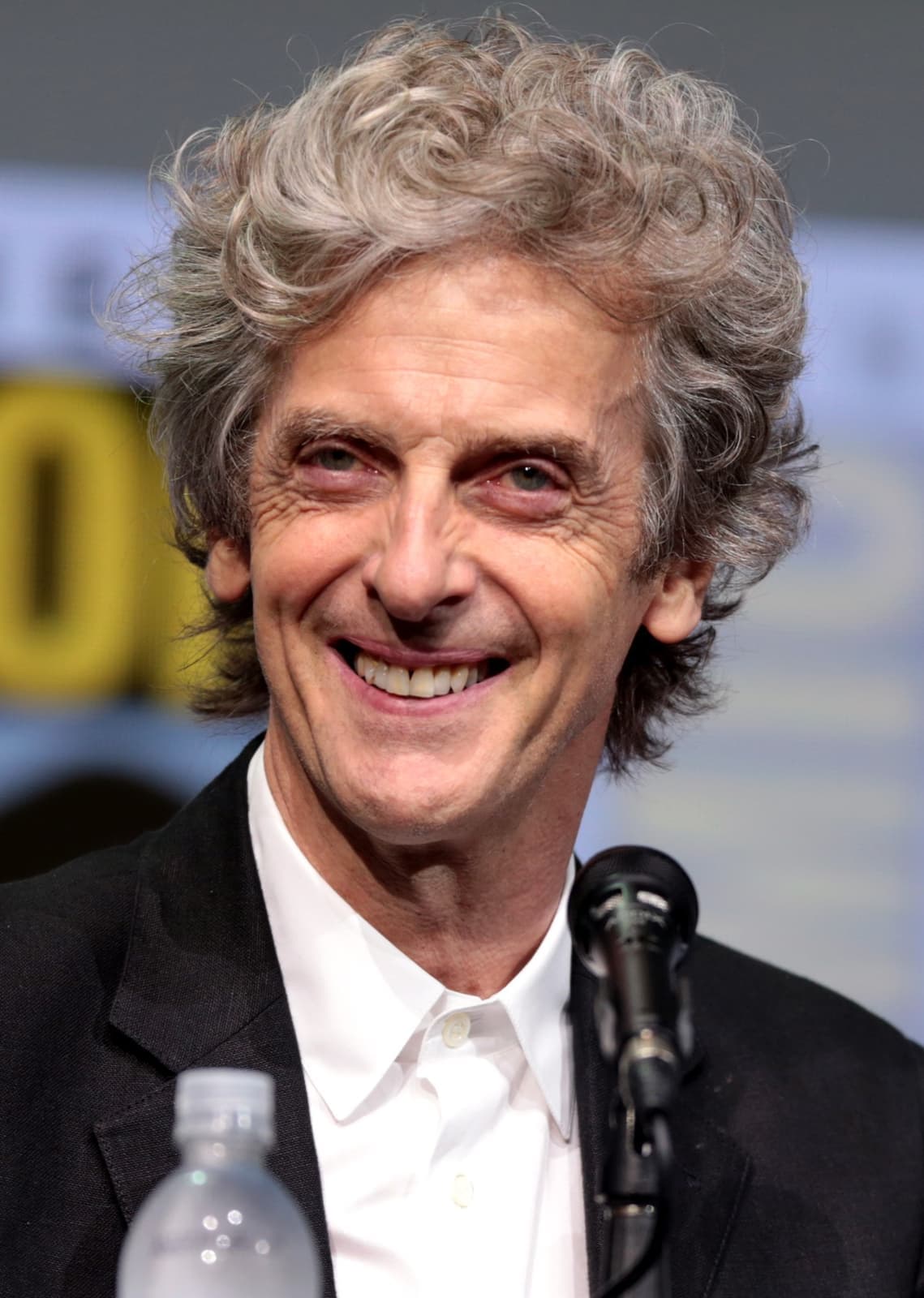 Peter Capaldi smiling, grey curls, at a panel microphone