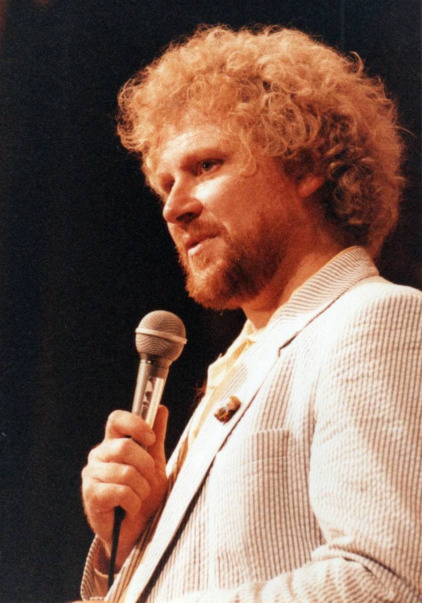 Colin Baker, with a mass of curly hair and a beard, speaking into a hand microphone