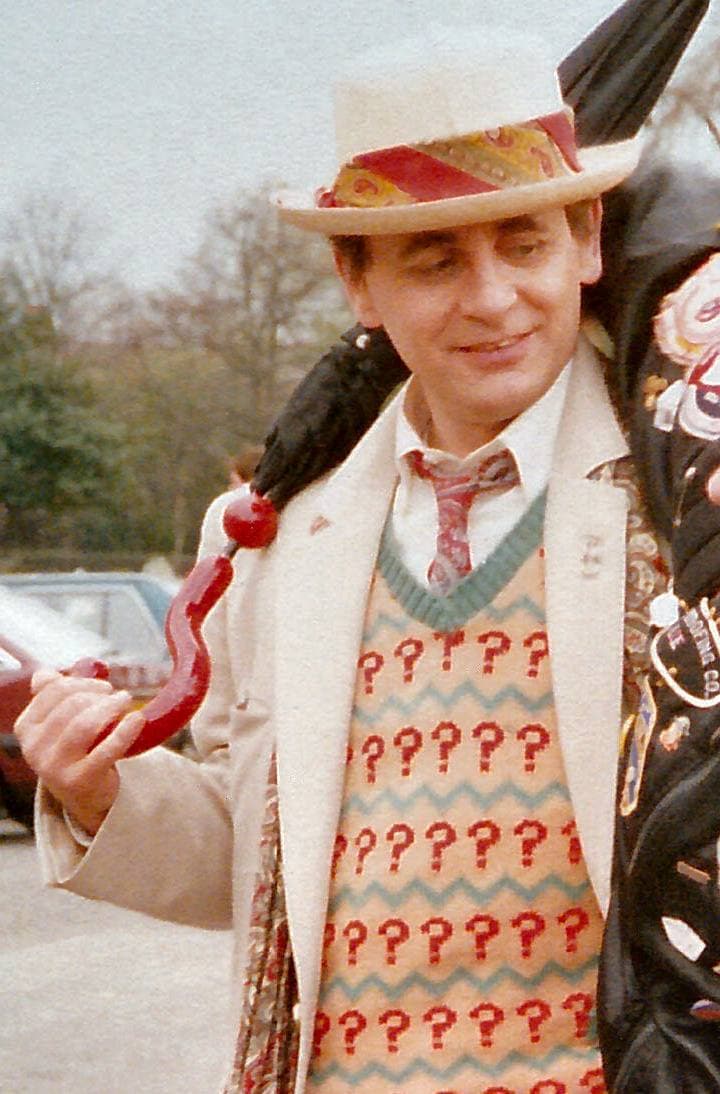 Sylvester McCoy in a straw hat and question-mark pullover, holding a red-handled umbrella
