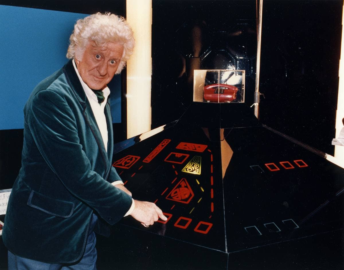 Jon Pertwee, white-haired, in a velvet jacket with a frilled shirt, his hand on a dark hexagonal console