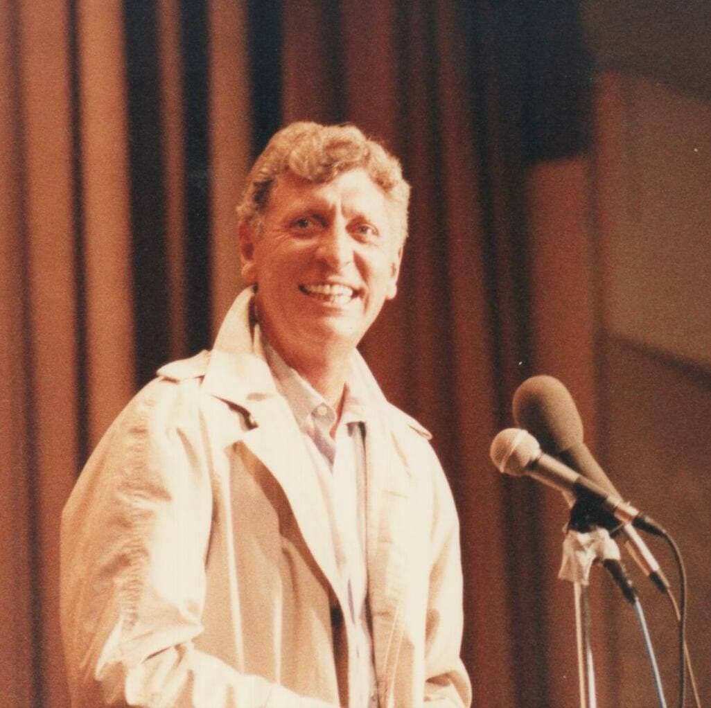 Tom Baker, curly-haired and grinning, in a pale raincoat at a microphone in front of a stage curtain