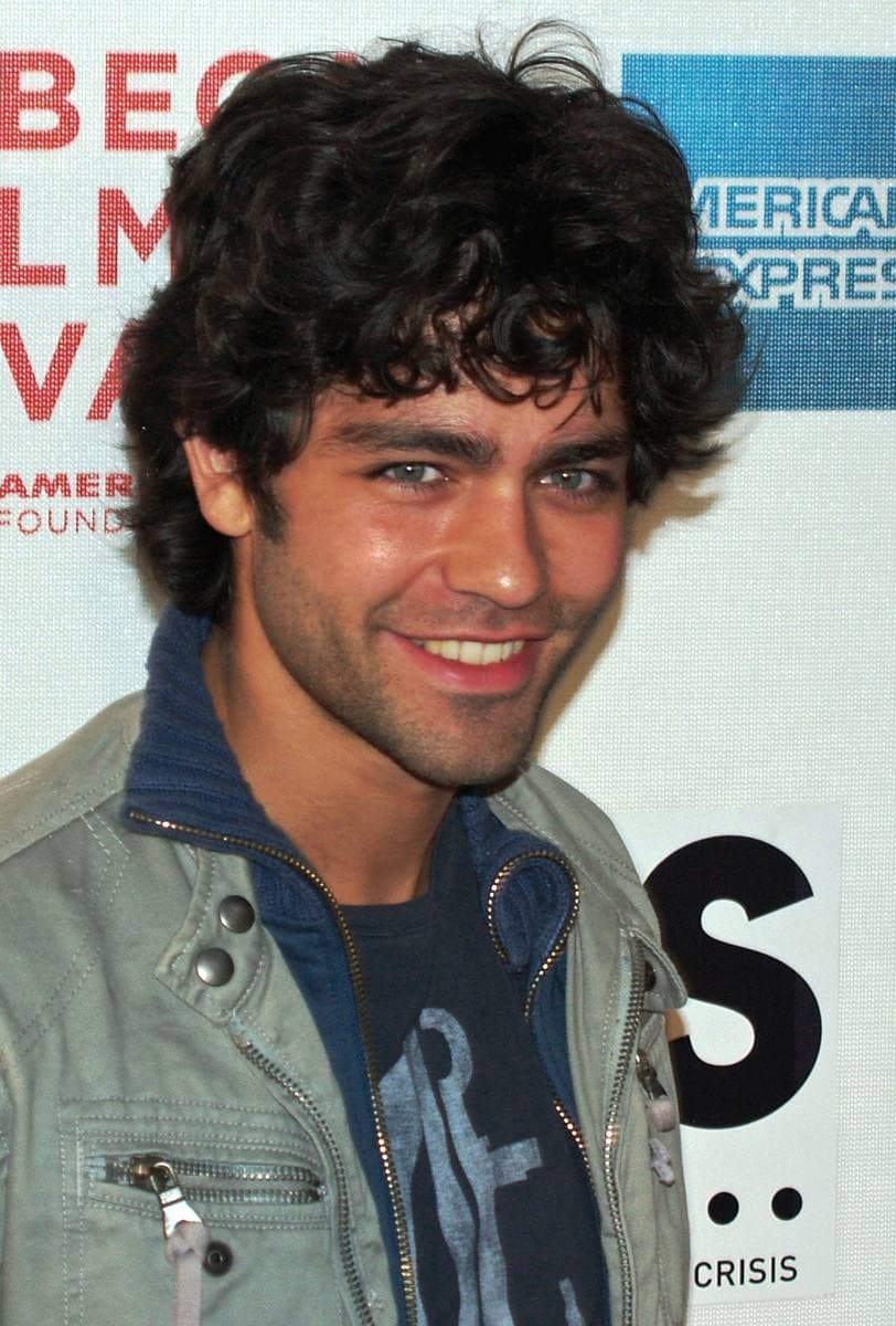 Adrian Grenier smiling, curly dark hair, in a pale green zipped jacket over a navy T-shirt, in front of a festival backdrop.