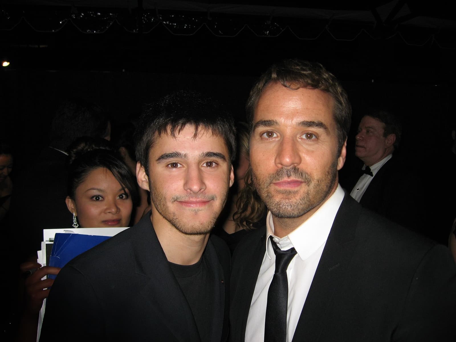 Jeremy Piven in a black suit and tie at a night event, with another man at left