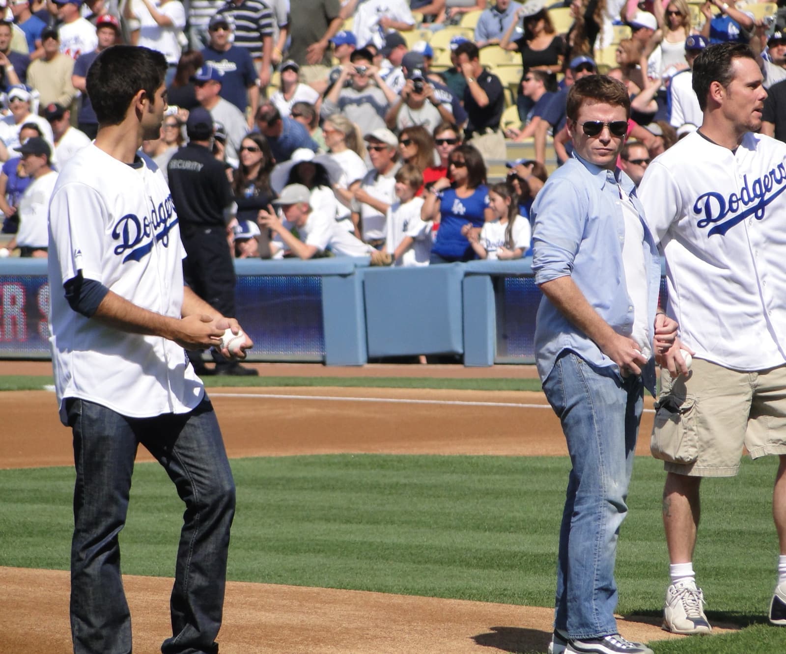 Three men on a baseball diamond, two in Dodgers shirts, crowd behind
