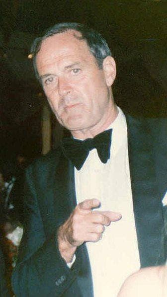 John Cleese in a dinner jacket and bow tie, pointing, against a dark background