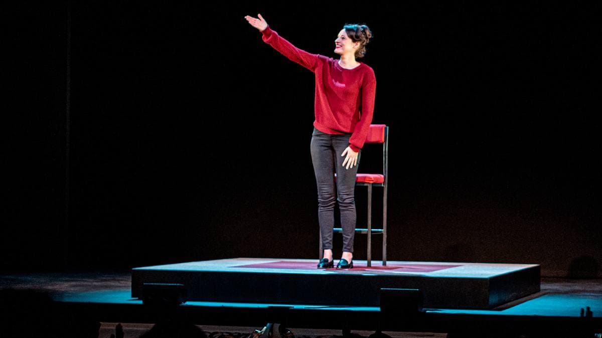 A woman in a red jumper and dark trousers stands on a small low stage beside a tall red chair, one arm raised, in a pool of light on a black stage