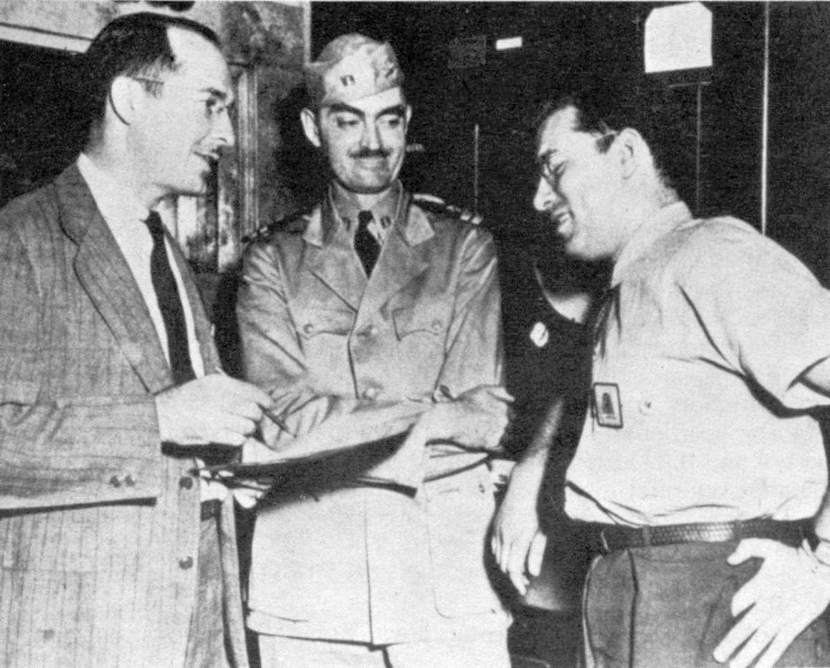 Three men in shirts and ties standing together in an office, the one on the right young and dark-haired
