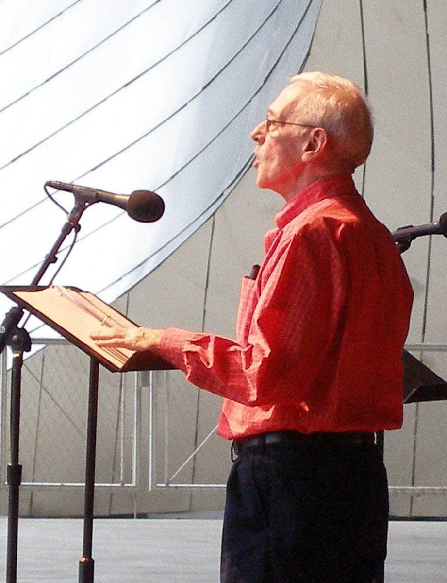 John Mahoney in a red shirt reading at a lectern on an outdoor stage