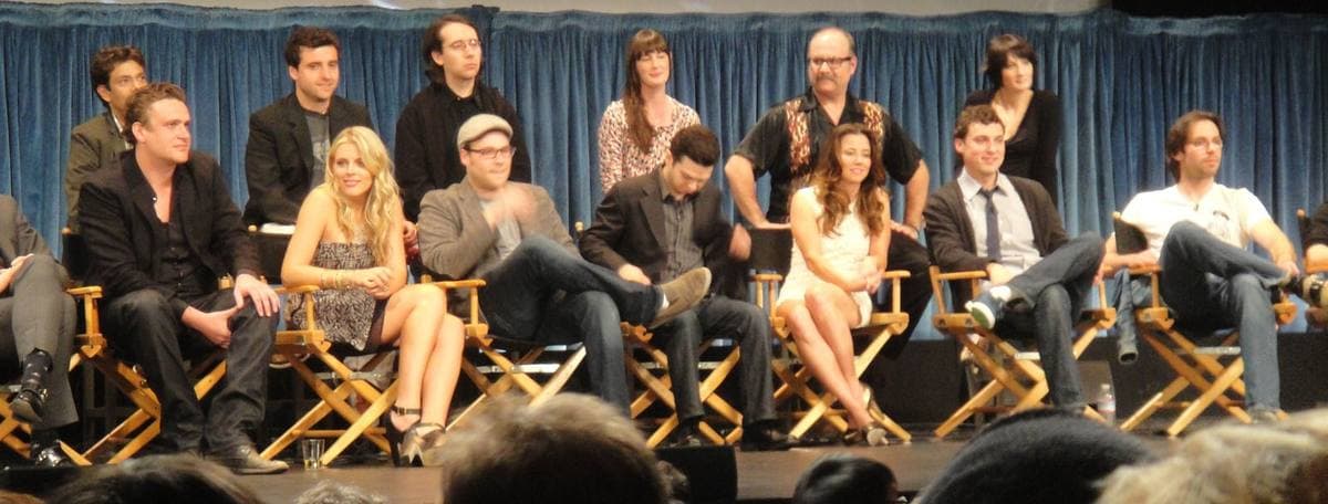 Thirteen people sitting and standing in two rows on a stage, smiling