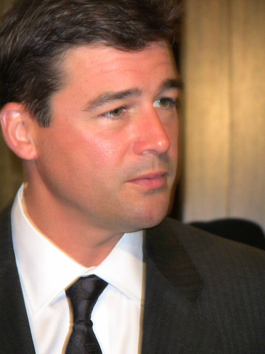 Kyle Chandler in a dark suit and tie, looking off to one side