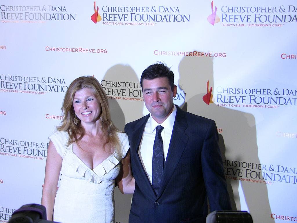 Connie Britton in a cream dress and Kyle Chandler in a dark suit smiling side by side in front of a sponsor wall