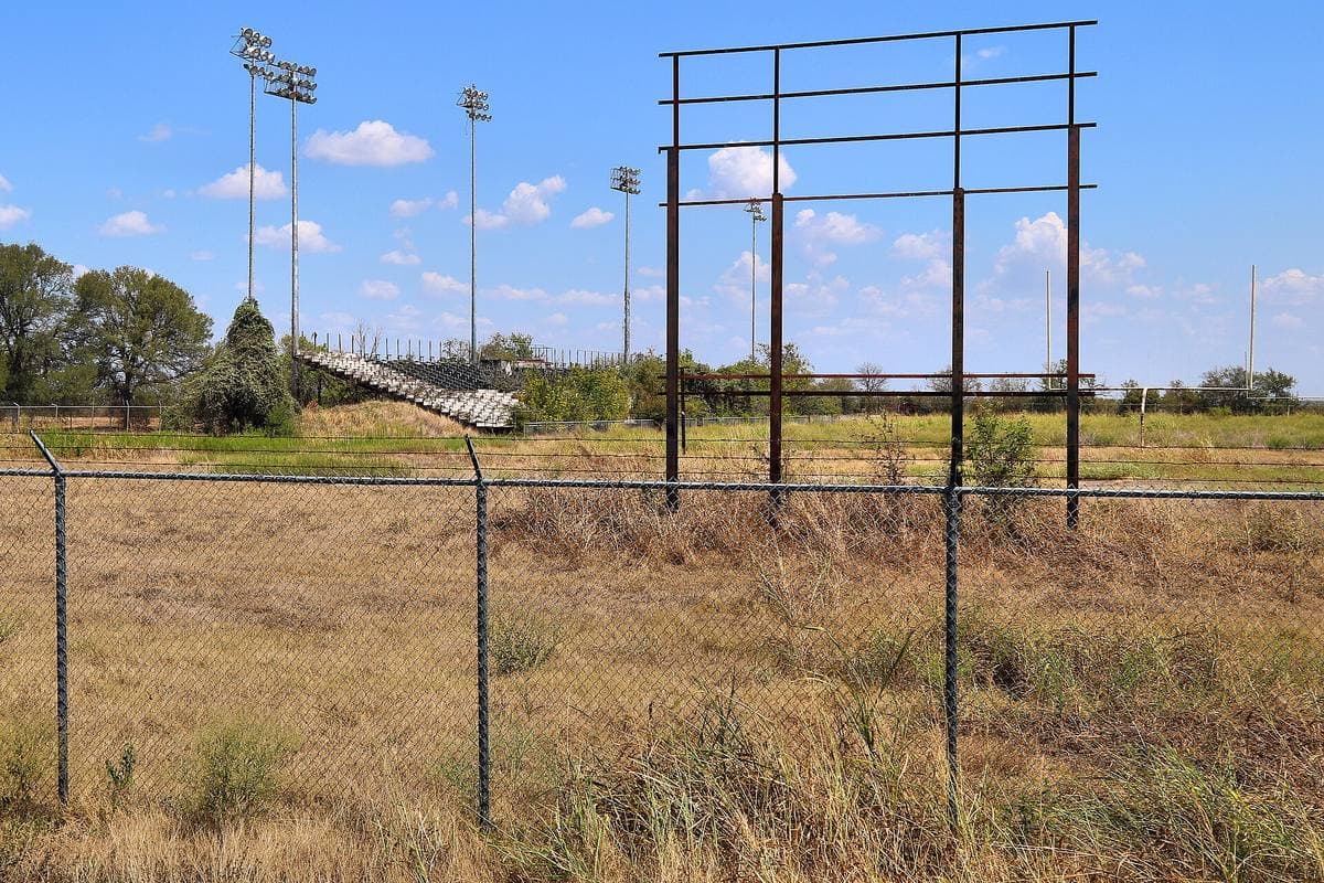 An overgrown football field behind a chain-link fence, with tall light towers and an empty steel frame against a blue Texas sky