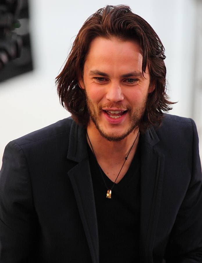 Taylor Kitsch with long hair and a dark jacket, laughing