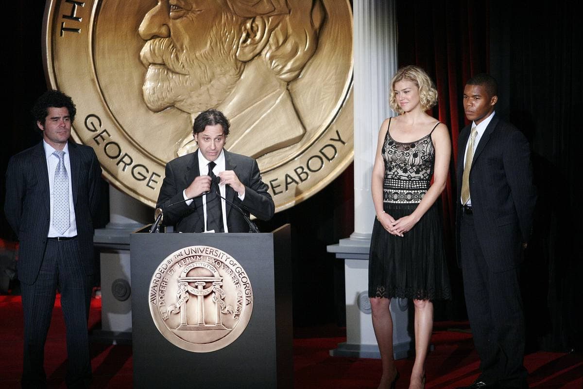 Four people on a stage in front of a giant gold Peabody medallion, one speaking at a lectern