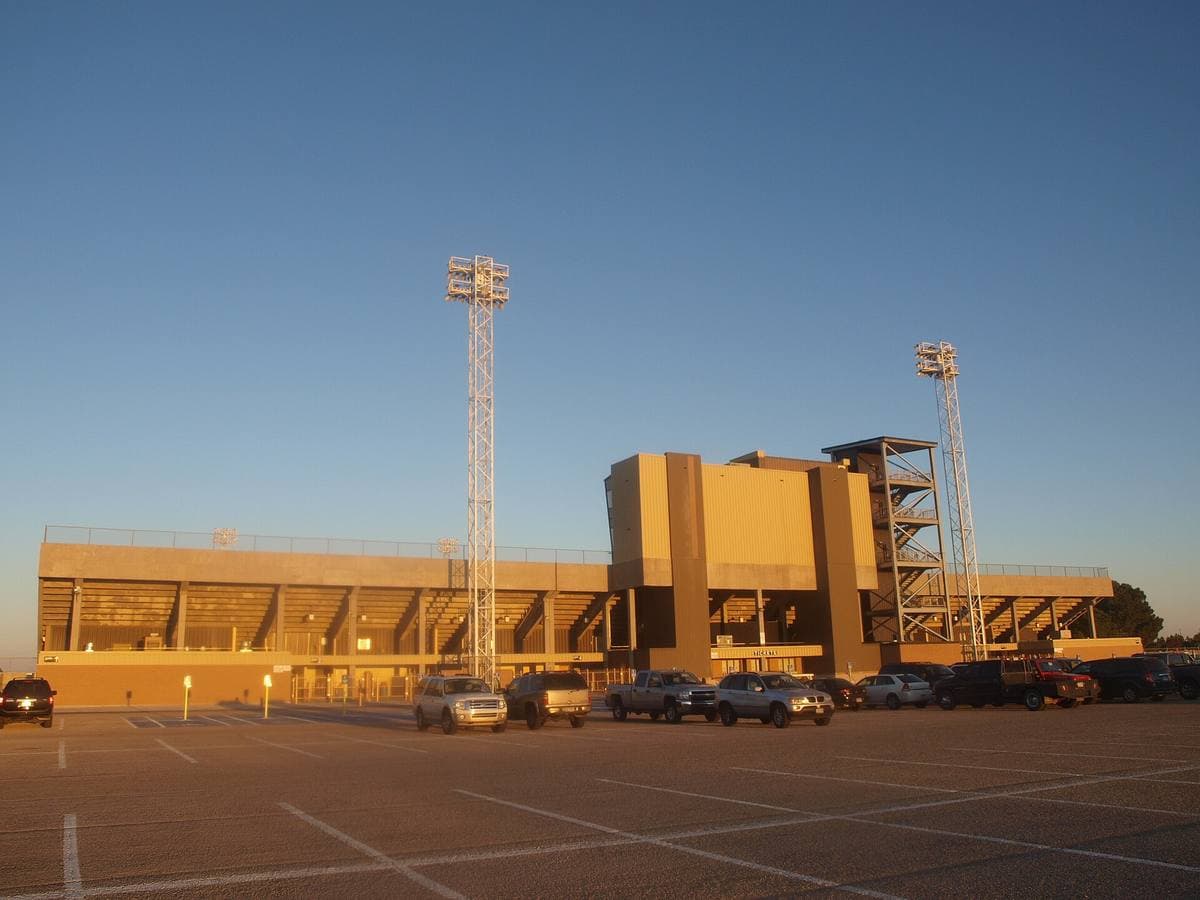 A tan concrete stadium with tall light towers glowing gold in late sun, seen across a car park