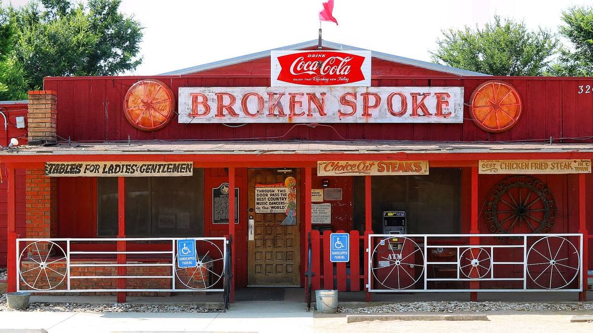 A red clapboard dance hall front with wagon-wheel railings and a hand-painted Broken Spoke sign