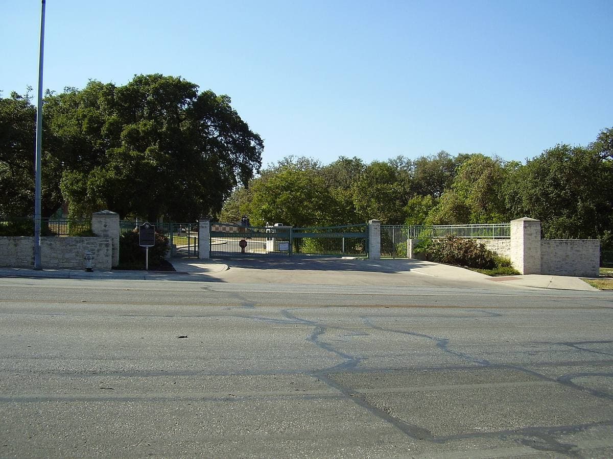 A stone gateway and low wall under live oaks beside a wide empty road