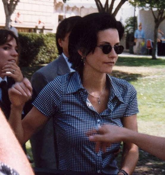 Courteney Cox in sunglasses and a check shirt, outdoors among photographers