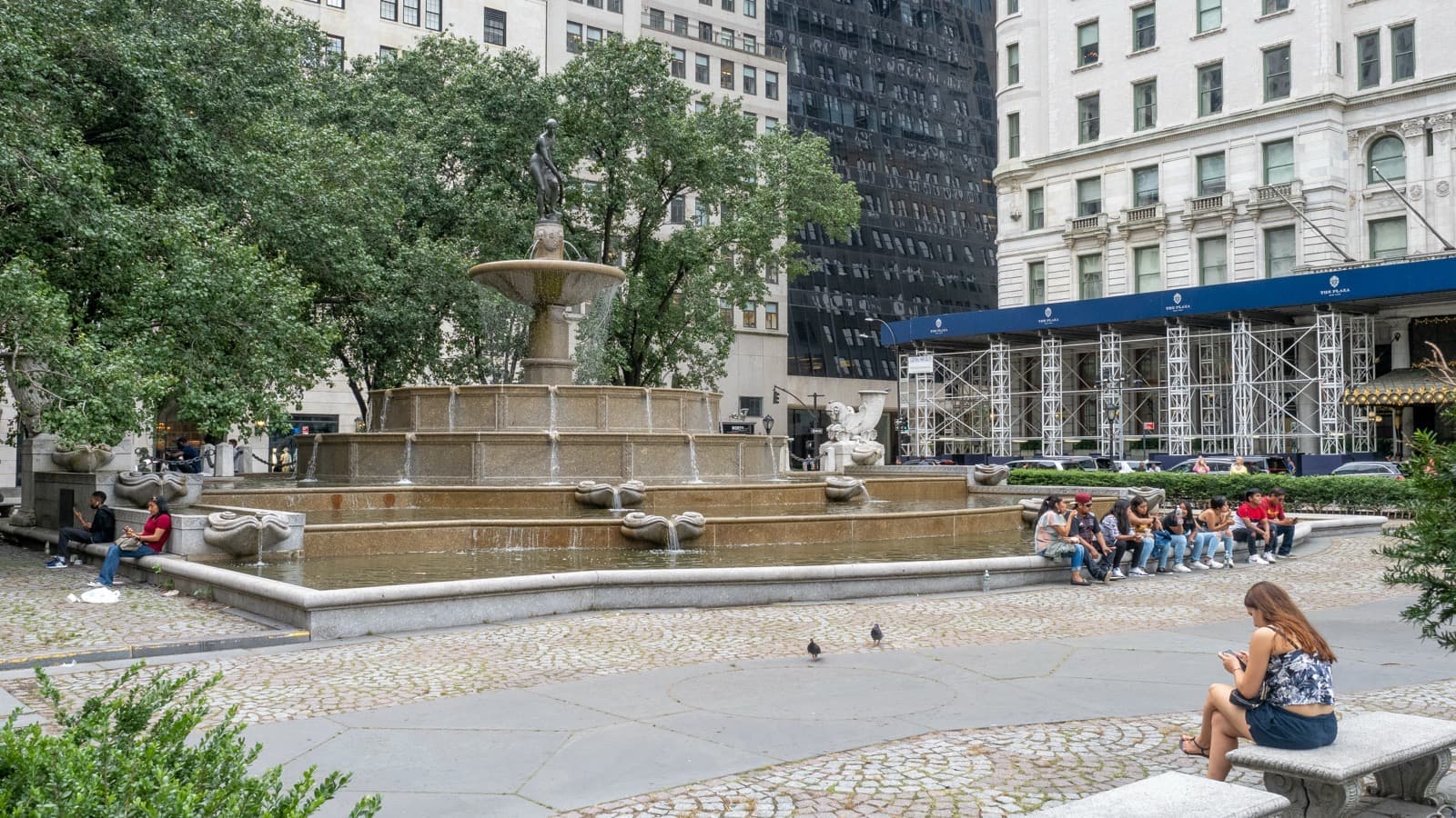 A tiered stone fountain topped by a bronze figure, with water falling between basins and the Plaza Hotel behind