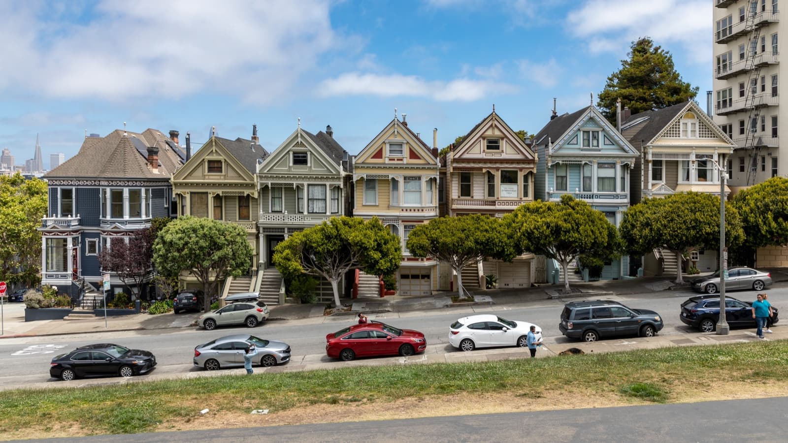 A grassy hillside with people sitting in the sun below a row of painted Victorian houses and the city skyline
