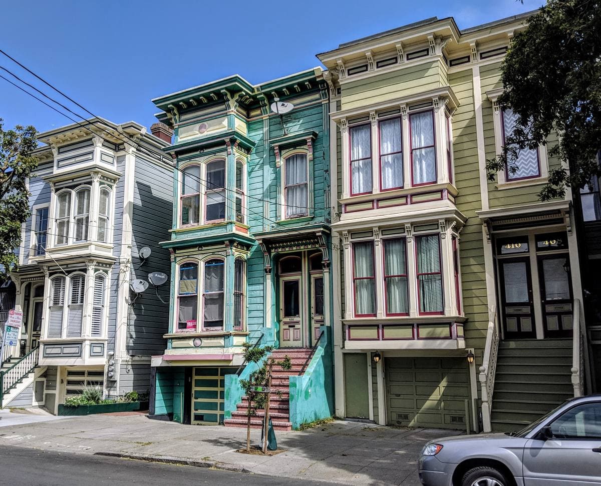 A row of painted Victorian houses with bay windows and front steps, green, cream and white, on a sunny street