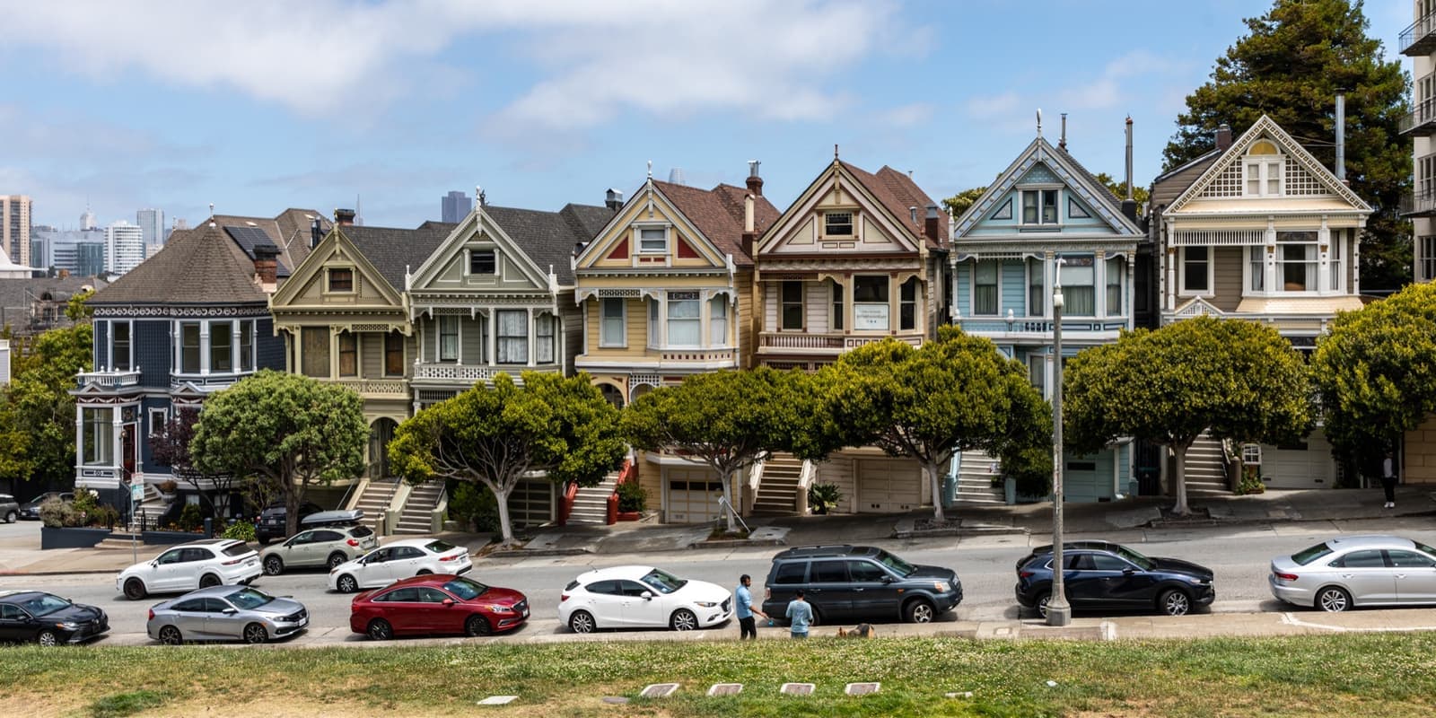 A row of pastel Victorian houses with steep gables facing a sloping lawn, downtown towers behind