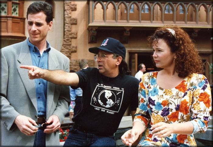 Bob Saget in a grey jacket, a man in a black cap and T-shirt pointing, and Gail Edwards with curly hair in a floral top, on a studio lot