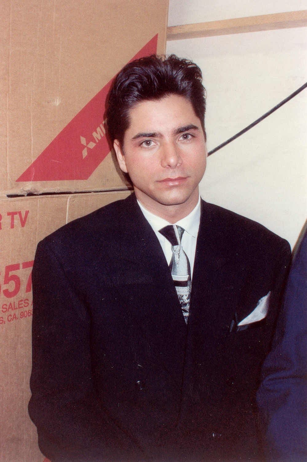 John Stamos smiling in a dark jacket, his hair swept back