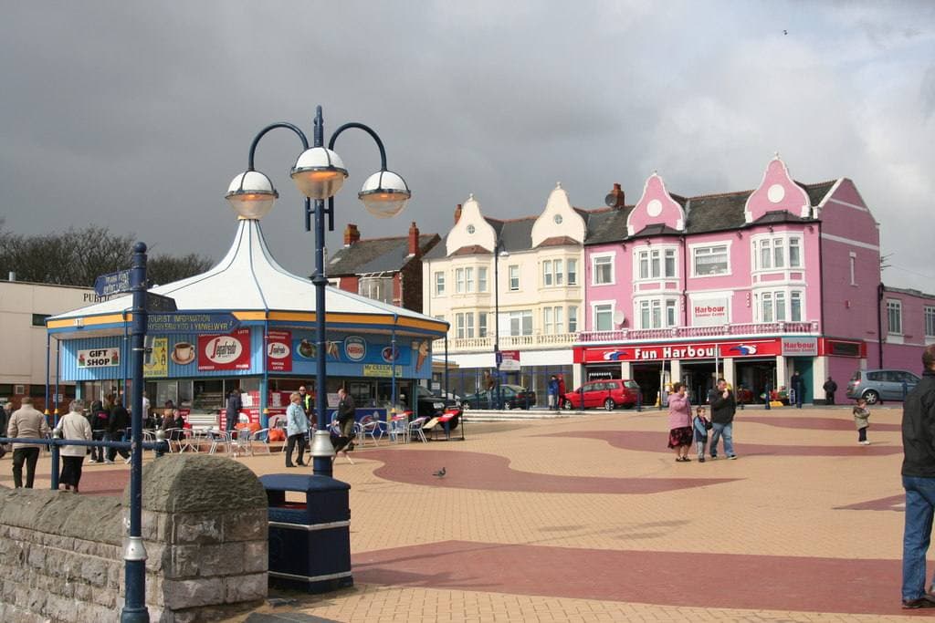 A paved seaside promenade with a carousel kiosk, lamp posts and a row of pink and cream arcade buildings