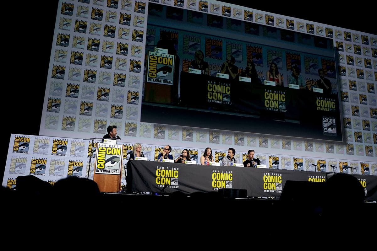 Six people seated at a long panel table on a stage, speaking into microphones