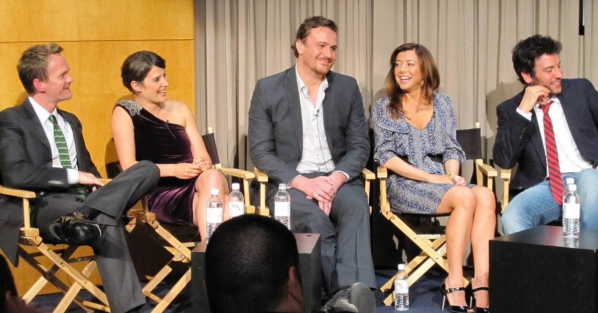Five actors seated in director’s chairs on a small stage, laughing