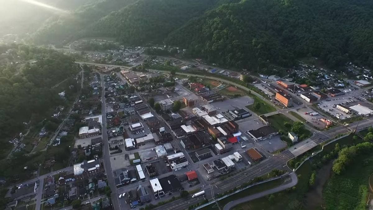 A small-town main street of brick buildings below wooded hills